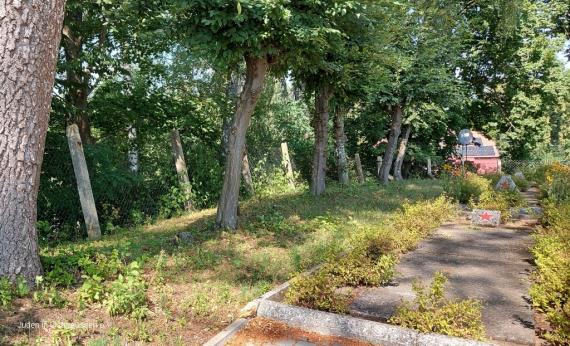 On the right concrete slabs with two short stelae with red stars. Diagonally in the center a strip of lawn, on it a row of trees, behind it a wire mesh fence with concrete pillars.