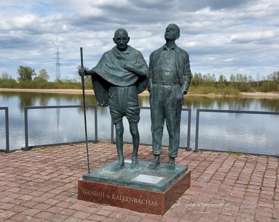 A monument with two metal figures, life-size men, the left one with staff, glasses and Indian clothes, the right one with European clothes and with sandals. Behind them is the water of a river.