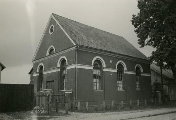 Black and white picture, a one-storey small house with long side facing the street with three windows, which are round at the top. The front side has two more windows of this type, in between there is a smaller door in the middle. To the street a very low fence, a sidewalk in front of it and some street with cobblestones.