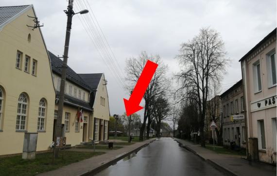 Color photo, empty street, in the foreground on the left the bright large two-story building with two gables to the street side and in between the building part that connects both gables. Behind it a gap in the building covered with lawn.