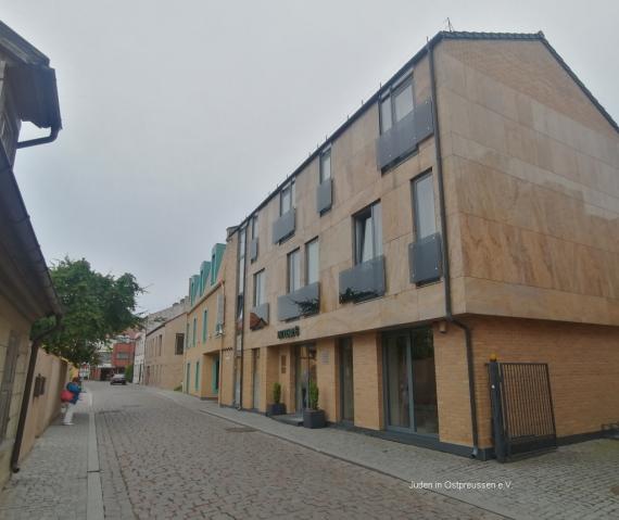 A yellowish, modern-looking building with a gable roof and three floors. In front of the windows dark glass attached as a balcony lattice. Behind it, a narrower building with turquoise window and door frames. In front of it a street with cobblestones.