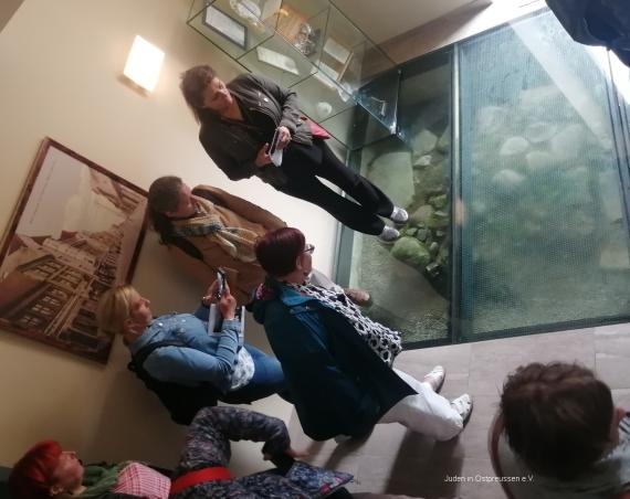 The picture shows diagonally from above in a room a group of people looking at a wall remnant under a glass plate embedded in the floor.