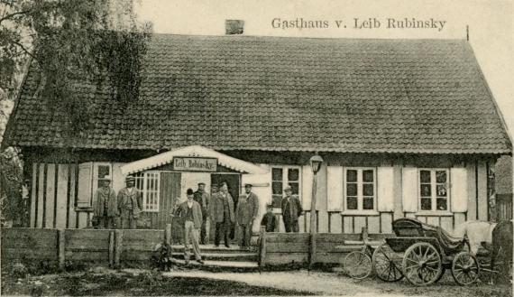 Long side of a one-story house with large roof. In front of it are more than 10 people. Above the entrance a sign with the name Keib Rubinsky. In front of it a low board fence, on the right there is a bicycle and a carriage with two horses.