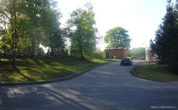Gedenkstätte für Juden aus Sensburg Narrow strip of grass in front of an existing cemetery fence, a large tree on it, a spur road in the middle with a parked vehicle, 2 small buildings on the right in the background.