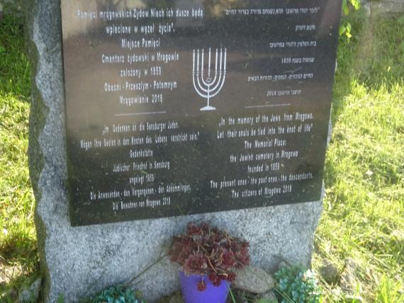 Dark gray stone slab depicting a seven-branched menorah candelabrum in the center, four text boxes with different languages and characters, mounted on gray stone stele, a small flower pot is at the bottom, green lawn background.