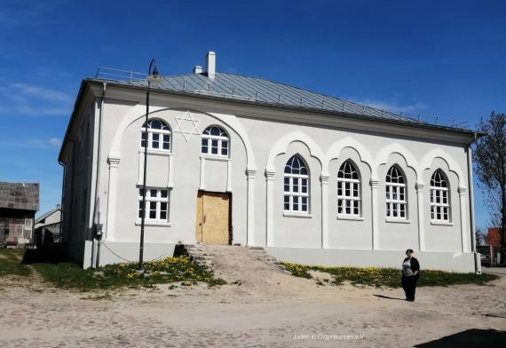 Gray two-story building with hipped roof. 4 windows on the right are long and pointed, on the left 2 areas with smaller windows and a door, this is made of preliminary wooden panels. In the foreground old cobblestone. To the door leads an eight-step staircase, which serves as a site ramp with backfill.