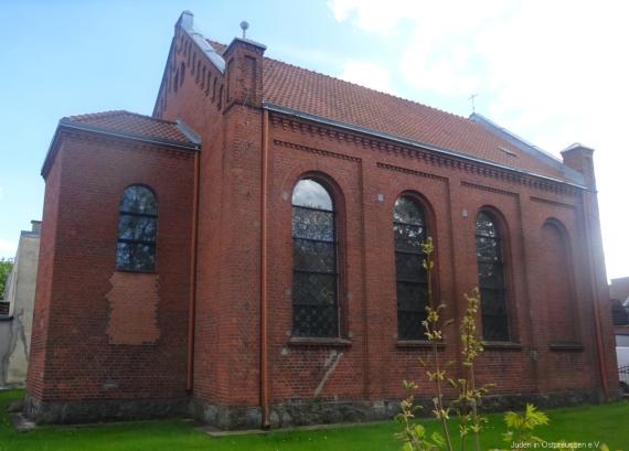 Seite und Rückseite der Synagoge. Hinten eine Eckige Absis mit bunten Glasfenster, Seite mit 4 hohen Fensternischen, drei mit Glasscheiben. Rote Dachziegeln auf dem Giebeldach. Umgeben von flachen grünen Rasen.