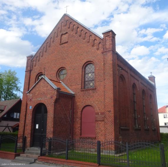 Reddish two-story brick building with small porch for the entrance. On the long sides 4 long windows with an arched termination at the top. The corners of the building end in small ornamental turrets. A small cross can be seen on the gable. In front of the building a black metal fence.