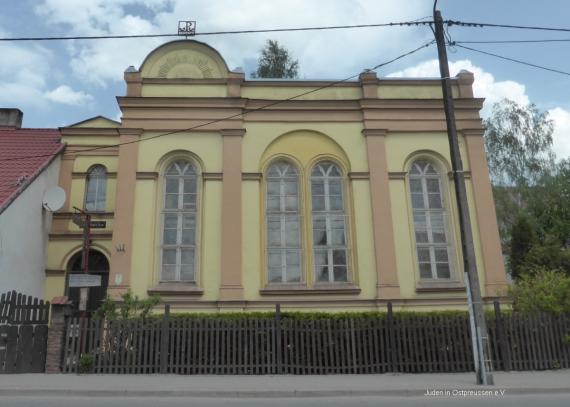 A yellowish plastered building with 4 long windows ending arched at the top. A roof is not visible. At the top of the facade is a small decorative arch. On the left side a brown entrance door is shifted a little bit to the rear. In front of the building is a picket fence, sidewalk and asphalt road.