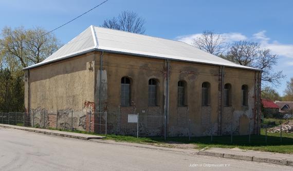 Building with six windows on the long side. Windowless front side facing the street. In front of the building a wire mesh fence. A glossy silver hipped roof.