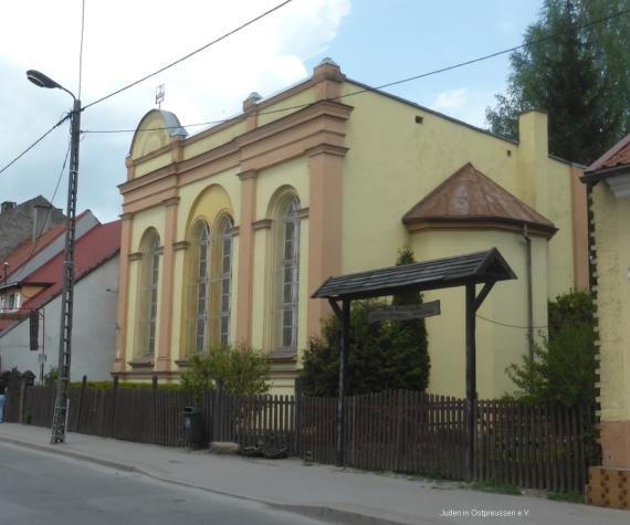 A yellowish building obliquely seen from the front. You can see a semicircular absis on the right side at the front of the building with a red tin roof. To the street side 4 large windows. In front of it a picket fence with an entrance gate that has a small roof with wooden shingles.