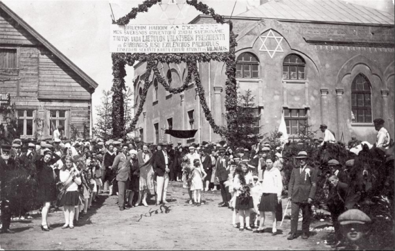 Im Hintergrund rechts ein großes Synagogengebäude, links eine Holzhaus, dazwischen ein Gerüst mit Blätter-Girlanden. oben ein Willkommens-Transparent in Litauisch mit hebräischer Grußformel für den litauischen Staatspräsidenten.   