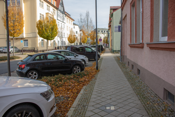 Standort Wohnung der Familie Ortenberger (Stolpersteine) Blick in die Bahnhofstraße vom Gehweg. Links sind andere Gebäude, die Straße und geparkte Fahrzeuge. Rechters sieht man das hellrote Wohnhaus der Familie Ortenberger, die heutige Ilmenauer Volkshochschule. Am unteren Bildrand sind die verlegten Stolpersteine zu erkennen.