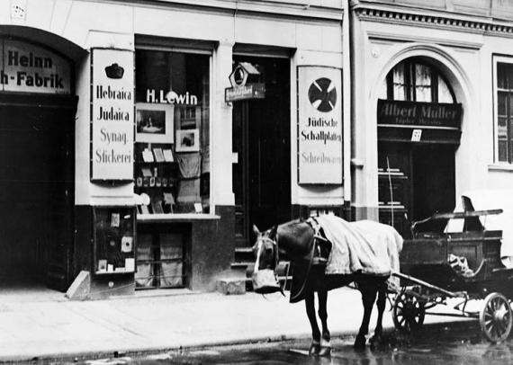 Ein Pferdekutsche vor einem Laden. Im Schaufenster sieht man gerahmte Bilder. Auf den Schildern steht "Hebräika Judaica Synog. Stickerei", "H. Lewin" und "Jüdische Schallplatten, Schreibwaren".