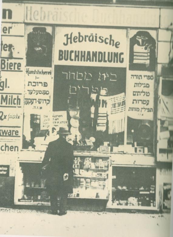 Das Schaufenster der Buchhandlung. Zu sehen im unscharfen sind viele verschiedene Waren, teilweise schwer zu erkennen, u.a. zwei Gebetschals und ein Chanukah-Leuchter. Auf Deutsch steht geschrieben "Hebräische Buchhandlung | H. Lewin". Auf Hebräisch und Jiddisch sieht man die Namen von vielen angeboteten religiösen Objekten, inkl. Torarollen.