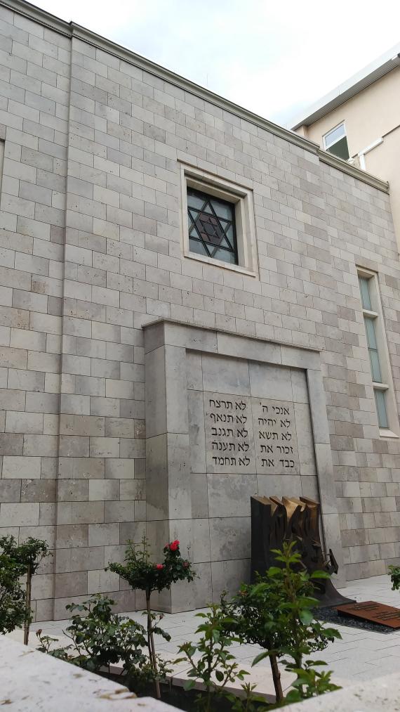 In the background you can see the facade of the Stuttgart synagogue made of light Tavertin stone. In the foreground red roses blooming in the garden. A bay window juts out from the facade, behind which is the synagogue's Torah shrine. On it can be seen the opening words of the Ten Commandments in Hebrew script. Above it is a window with a Star of David made of colored glass, through which the light of the sun shines during morning prayers and beautifully illuminates the interior of the Stuttgart synagogue.