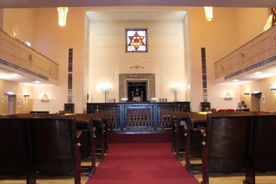 In the foreground you can see the lectern for the Torah reading (Bima), to the right and left of it the rows of seats. In the background, the Torah shrine (Aron HaKodesh), covered with a blue embroidered curtain (parochet). Above, the morning sun shines through the colorful window with the Star of David. Above, two of the four candelabras from the 1950s can be seen, as well as the gallery surrounding the right and left sides and the back.