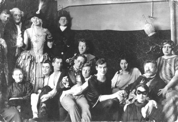 black and white shot of a group of about 15 people standing and sitting looking at the camera or looking at each other. They are dressed up in expensive dresses, wigs and masks in the style of the 1920s. Many are laughing or have their arm around each other.