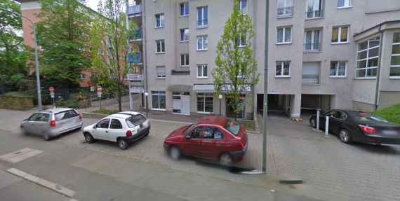 Es wurde von der Straße aus fotografiert. Autos stehen vor einem mehrstöckigem Wohnhaus. im erdgeschoss sind Fenster und eine Markise mit grün-weißen Streifen zu erkennen.