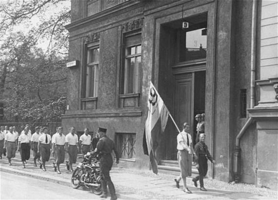 black and white shot, building facade of a bourgeois house with large doors, to see are in line marching white blouses wearing women and men in uniform. A young man with flag passes by, probably with swastika