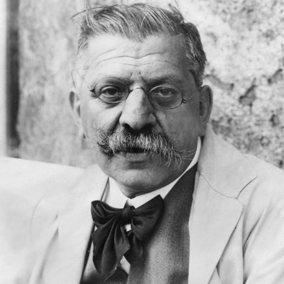 You can see a black and white photo of Magnus Hirschfeld. You can see round glasses and his moustache. He is wearing a jacket with a black bowtie.