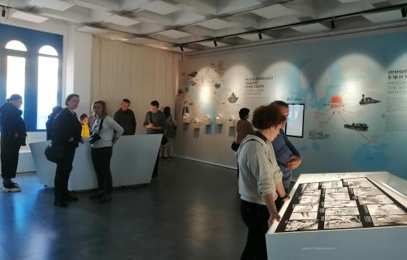 People In the museum hall, they stand at illuminated tables with comic-like representations, behind the wall kept in light construction with showcases, graphics and a screen.