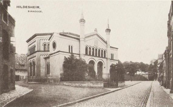 The view falls on the white synagogue whose entrance arches are flanked by two pointed towers. In front of it there is a small garden. The building has round-arched windows on all sides.