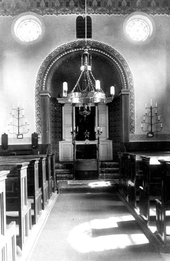 Synagoge, Inneres von Westen, mit Blick auf den Thoraschrein, links und rechts sieht man hölzerne Sitzreihen, das Foto ist aus zentraler Position aufgenommen. Beiderseits des Schreins sieht man an der Wand angebrachte Chanukkiot.