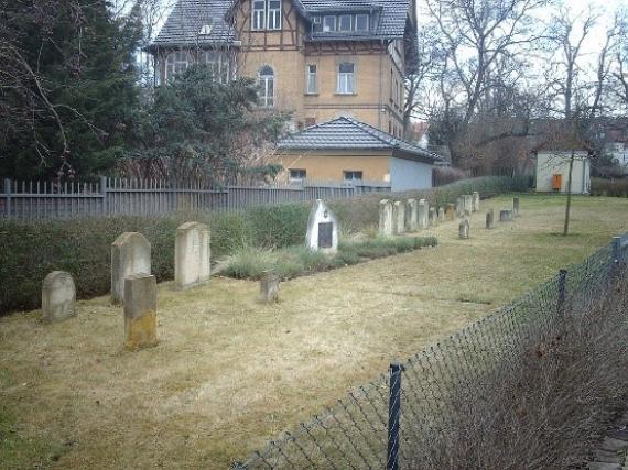 A strip of lawn with several Jewish gravestones lined up in a row. In the background are a villa and trees.