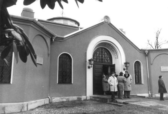 Black and white photo of a flat building with a pointed gable and round windows. People in winter coats are standing in front of the building.