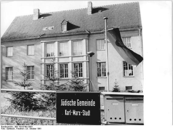 Jüdisches Gemeindehaus Chemnitz (Stollberger Straße 28), 1961 (Jahr der Einweihung) Black and white photo of a large two-story building with a garden. A GDR flag flies on a flagpole in front of the building. A sign reading "Jewish Community Karl-Marx-Stadt" hangs on the fence in front of the building.