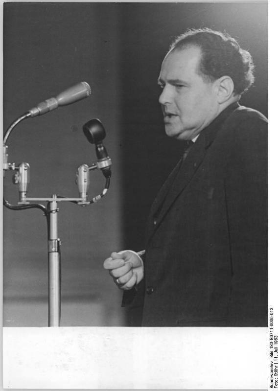 Black and white photo of an elegantly dressed man in profile speaking into a microphone.