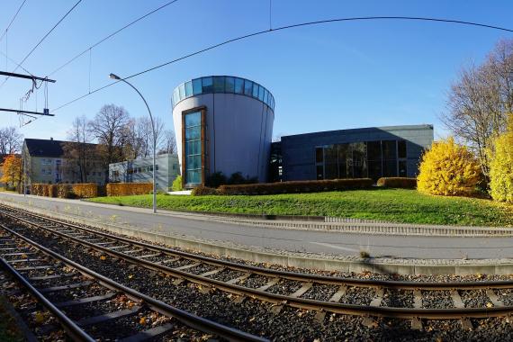 Die Neue Synagoge in Chemnitz an der Stollberger Straße, 2015 Cylindrical modern building with a glass window wall on one side. In its surroundings are other modern buildings and green lawns. Streetcar tracks run in the foreground.