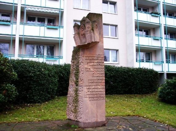 Memorial column made of reddish stone. On it in block letters the inscription: This was the site of the synagogue consecrated by Rabbi Dr. Mühlfelder in 1899. It was reduced to rubble by fascist arsonists on the night of the pogrom on November 9, 1938. In the background is a modern apartment building.