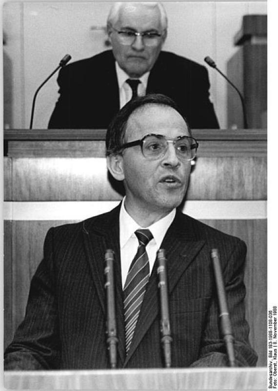 Black and white photo of a man with glasses and a suit. He is standing in a plenary chamber and speaking into a microphone. Behind him, further up, sits another man in a suit.