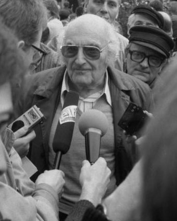 Black and white photo of an older man with sunglasses and coat in a crowd of people. Microphones are held out to him from all sides.