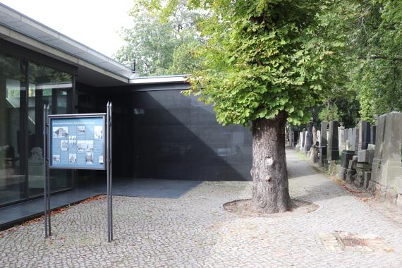 Vorplatz des Friedhofs mit Infotafel und Baum, rechts erste Grabsteine