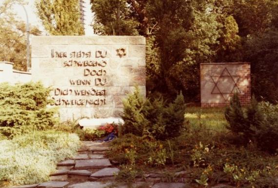 Gedenkstein mit Aufschrift umgeben von Büschen und einem Weg