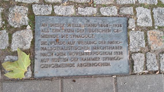 Auf dem Bild ist eine metallene Gedenktafel mit Reliefschrift im Boden zu sehen. Die Tafel erinnert an die ehemalige Synagoge in Hamm.