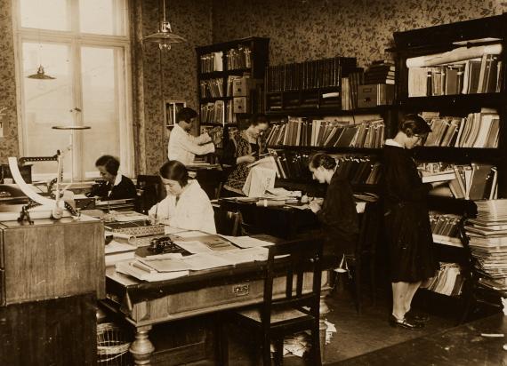 Women work at desks, search through bookshelves