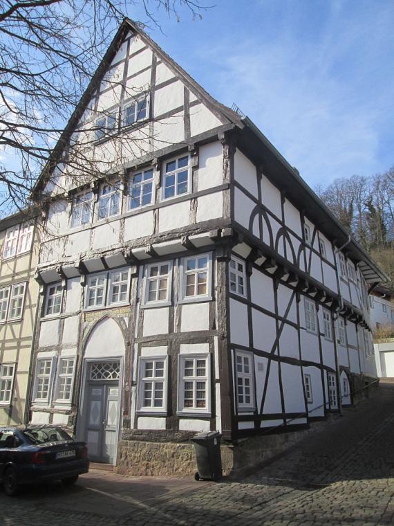 The picture shows a three-storey half-timbered house from the front and the side. The sky is blue and a bare tree juts into the side of the picture.