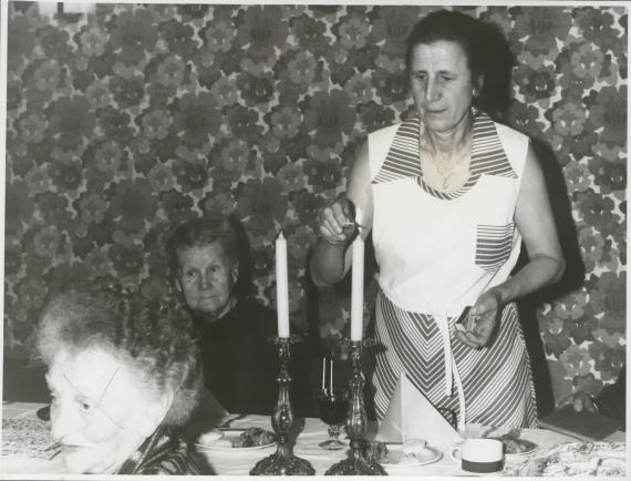 Three women at a table, one woman standing lights two candles