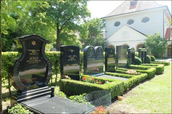 Seitliche Ansicht auf einen Teil des Friedhofsfelds mit Grabsteinen. Dahinter ein Haus.