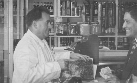 Two people talking to each other at the counter, numerous items of various delicacies for sale in the background