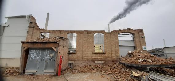 Farbfoto einer Fabrikruine, die abgerissen wird, mit zahlreichen Backsteinen am Boden liegend, im Hintergrund eine neue Fabrik