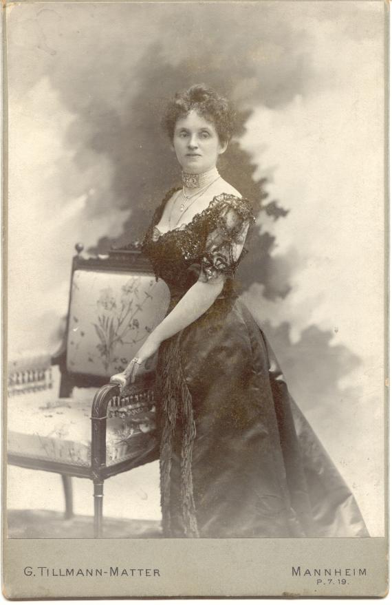 Studio shot of a woman standing in a dress, leaning against a chair, looking towards the camera