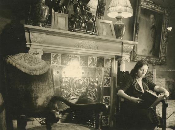 Black and white photograph of a young woman sitting on a chair in front of a fireplace and engrossed in a book