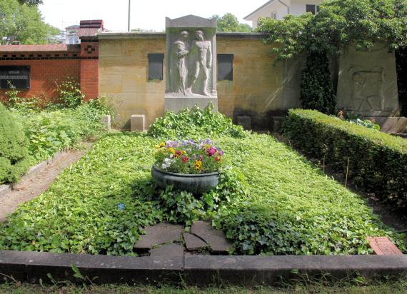 Fotografie einer Grabanlage mit üppigem Efeu, Blumenbeplanzung und Denkmal