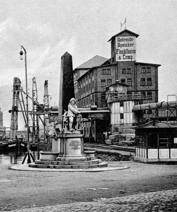 Black and white photograph of a detached brick warehouse building, with cranes and ships in the background, an obelisk and a seafaring monument in front of it