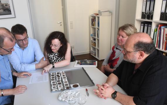 Several people sitting at a table, bent over a support plate with various coins and medals